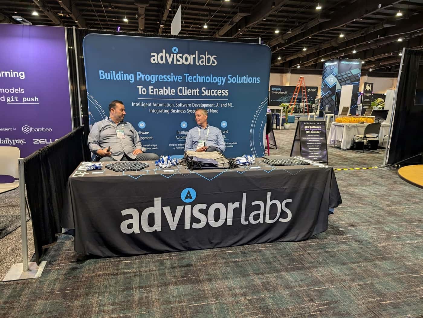 AdvisorLabs stand at a show: a curved backdrop headed Building progressive technology solutions to enable client success and a black AdvisorLabs table cover