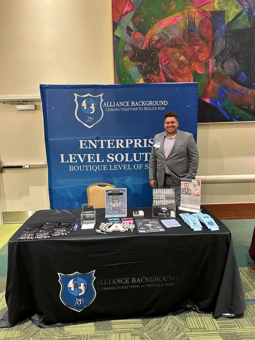 Alliance Background stand at a show: a blue backdrop with the shield logo and a black table cover with the same logo